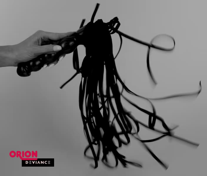 Fotografía en blanco y negro de una mano blandiendo un flogger negro con mango texturizado, con las tiras en movimiento