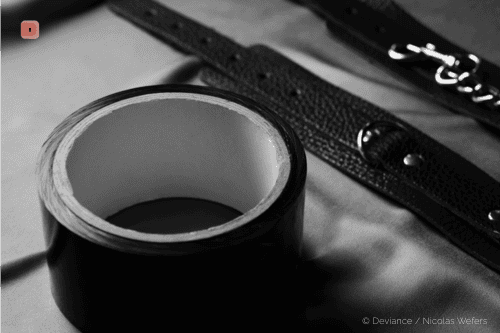 Primer plano en blanco y negro de un rollo de cinta de bondage negra, con esposas de cuero detrás