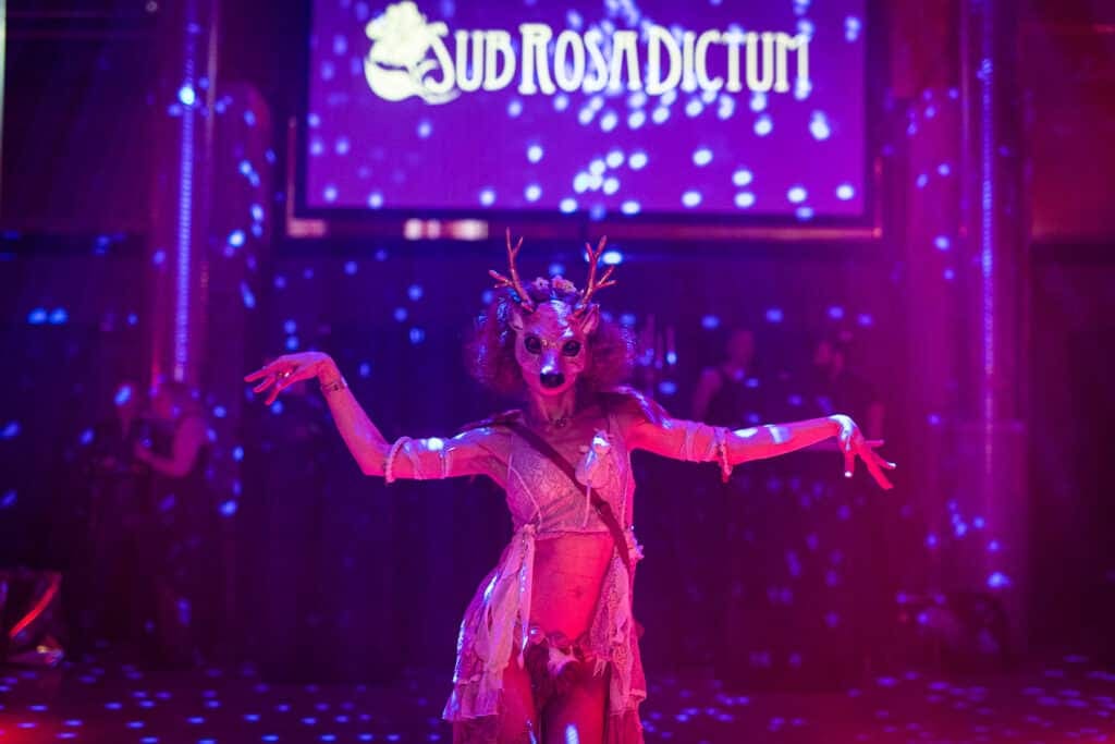 Artista con máscara de ciervo y cuernos baila con los brazos extendidos ante una pantalla con el rótulo «Sub Rosa Dictum»