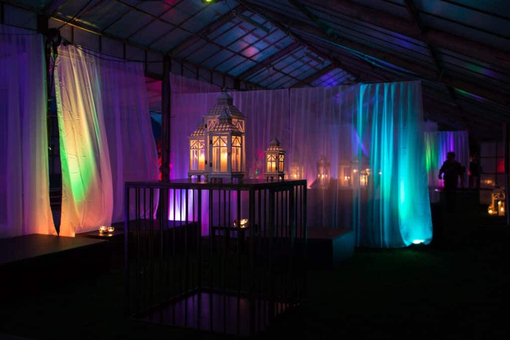 Zona de carpa de una fiesta fetichista iluminada con colores, cortinas blancas, grandes farolillos y una jaula metálica