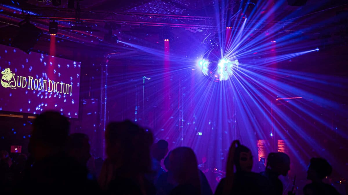 Pista de baile de una fiesta fetichista bajo una bola de espejos, con una pantalla que muestra «Sub Rosa Dictum» a la izquierda