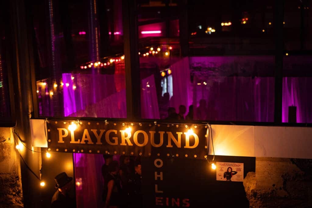 Cartel iluminado «Playground» con bombillas sobre la entrada a una zona de juego, con cortinas iluminadas en violeta detrás