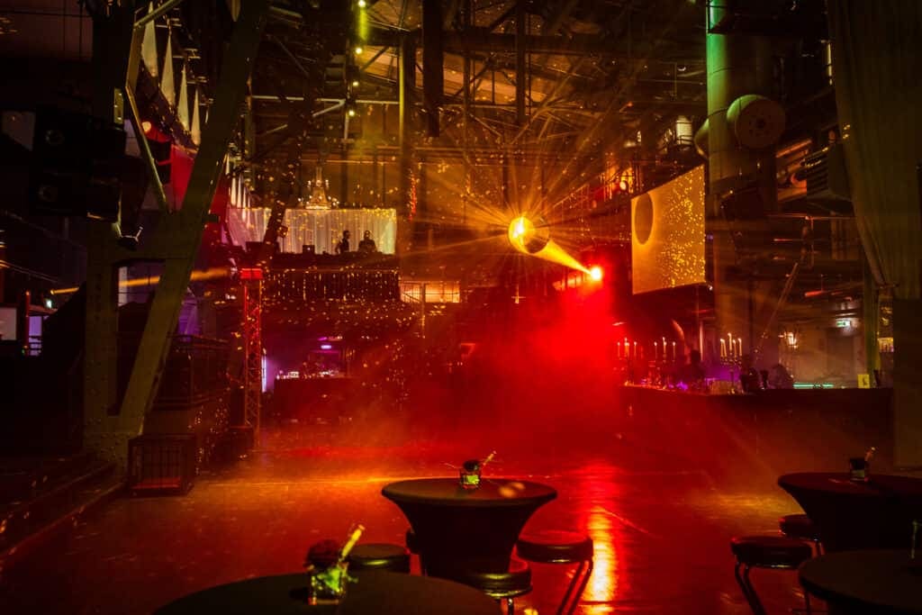 Sala vacía de una fiesta fetichista antes de abrir, iluminada en rojo y naranja cálidos, con mesas altas y cubiteras de champán