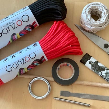 Material para el flogger DIY sobre una mesa de madera: paracord negro y rojo, anilla metálica, cinta aislante, hilo, tijeras, mechero, varilla de madera y herramientas