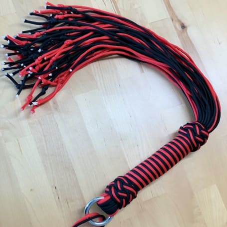 El flogger DIY de paracord negro y rojo terminado, con mango trenzado y anilla metálica, sobre una mesa de madera