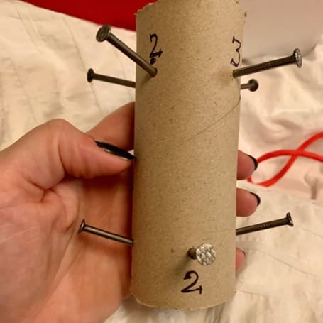 Una mano sostiene un tubo de cartón con clavos numerados clavados como guía para trenzar