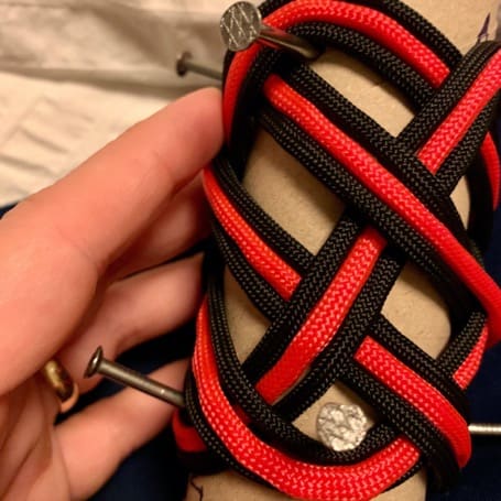 Paracord negro y rojo trenzado sobre los clavos del tubo de cartón formando un patrón