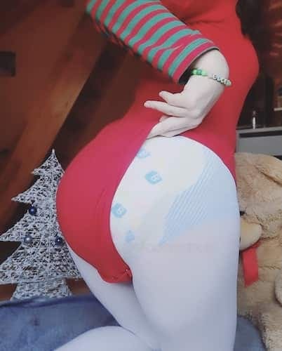 Persona con top rojo de mangas rayadas y un pañal estampado, junto a un osito de peluche y un árbol de Navidad plateado