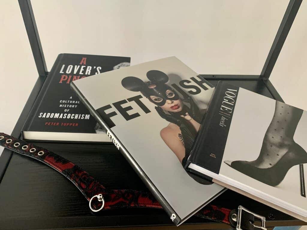 Libros ilustrados sobre fetichismo y sadomasoquismo en un estante negro — «Fetish», «A Lover's Pinch» y «Vogue Essentials: Heels» — junto a un collar rojo y negro