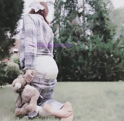 Persona con camisa de cuadros arrodillada descalza en un jardín, con pañal y un conejo de peluche en brazos