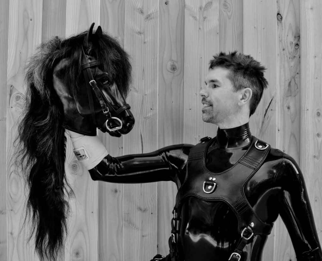 Fotografía en blanco y negro de una persona con traje de látex que sostiene sonriendo una máscara de caballo con crin y brida