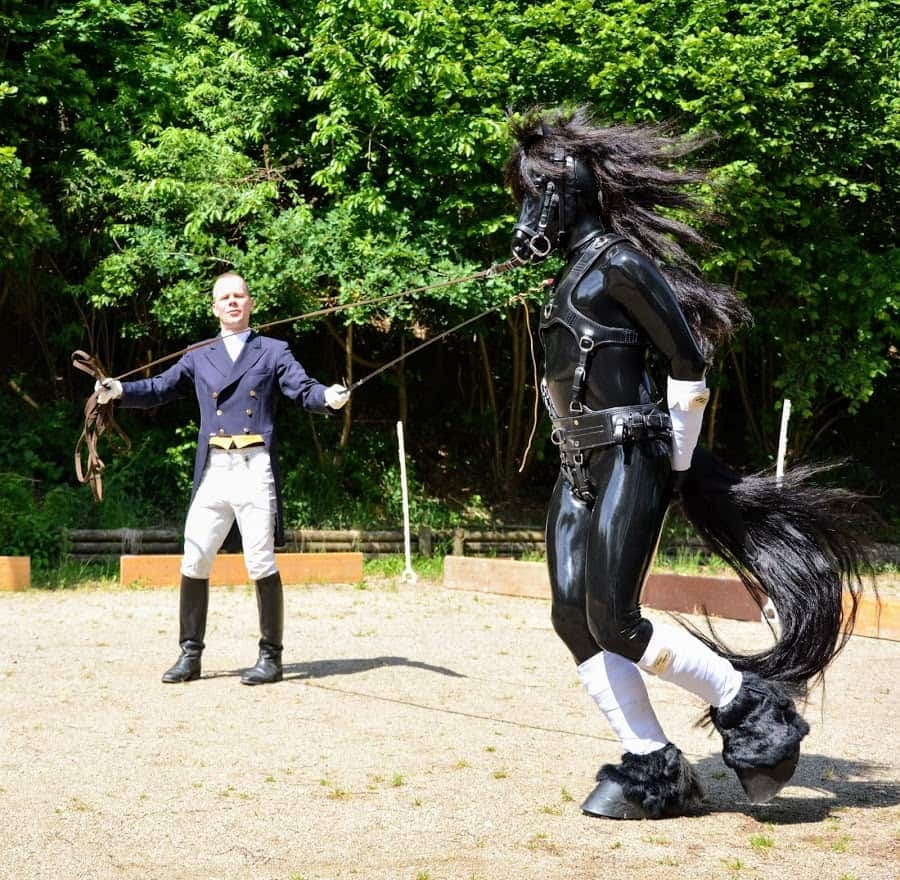Ponyplayer con traje de látex, máscara de caballo y penacho trota a la cuerda mientras un entrenador con ropa de monta sostiene una fusta en el centro