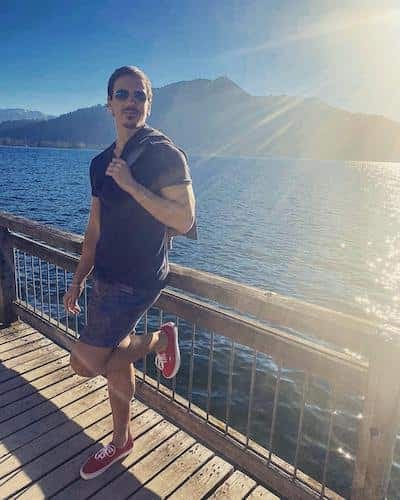 Persona con gafas de sol y camiseta oscura a contraluz sobre un embarcadero de madera junto a un lago de montaña