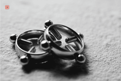 Primer plano en blanco y negro de dos pinzas para pezones circulares de metal con bolas en el borde y tornillos de ajuste interiores