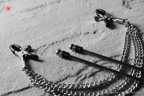 Fotografía en blanco y negro de varias pinzas para pezones sobre piedra: pinzas con tornillo regulable y cadenas colgantes, y dos pinzas de barra
