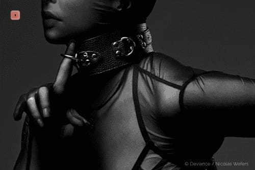 Primer plano en blanco y negro de una persona con un collar ancho de cuero granulado con varias anillas en O, llevándose un dedo a los labios