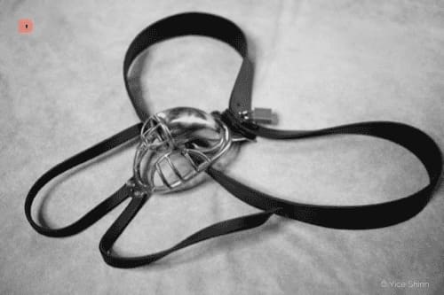 Fotografía en blanco y negro de una jaula de castidad metálica con correas negras y candado