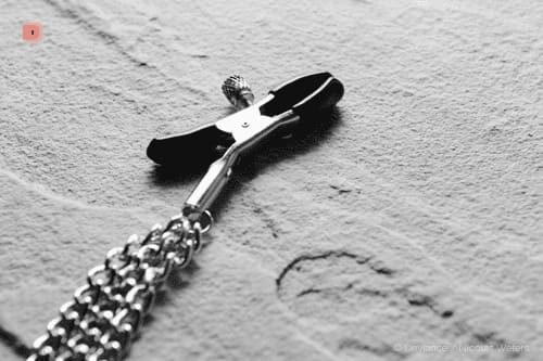 Primer plano en blanco y negro de una pinza para pezones con cadena colgante sobre arena clara