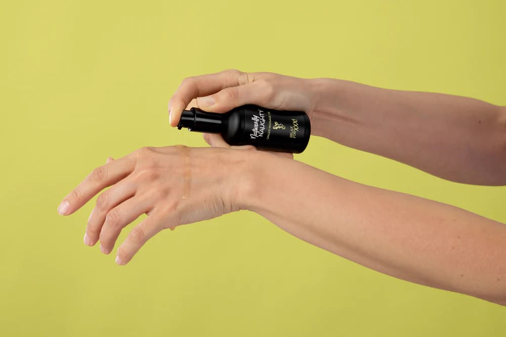 Dos manos aplican «Naturally Naughty Ginger Massage Oil» del frasco negro sobre el dorso de la mano, sobre fondo verde