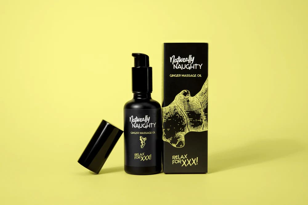 Frasco negro con dosificador de «Naturally Naughty Ginger Massage Oil» junto a su caja sobre fondo amarillo