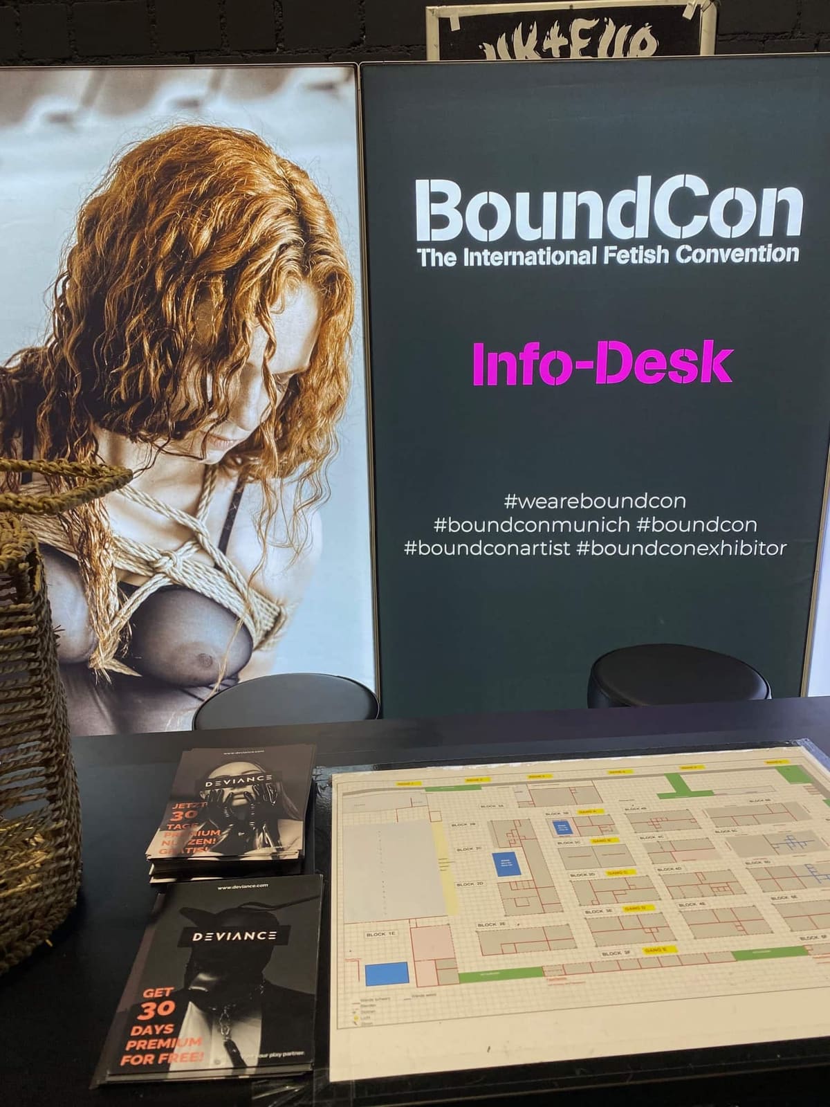Mostrador de información de BoundCon con un plano de la feria y folletos de Deviance, con un cartel de una atadura shibari detrás