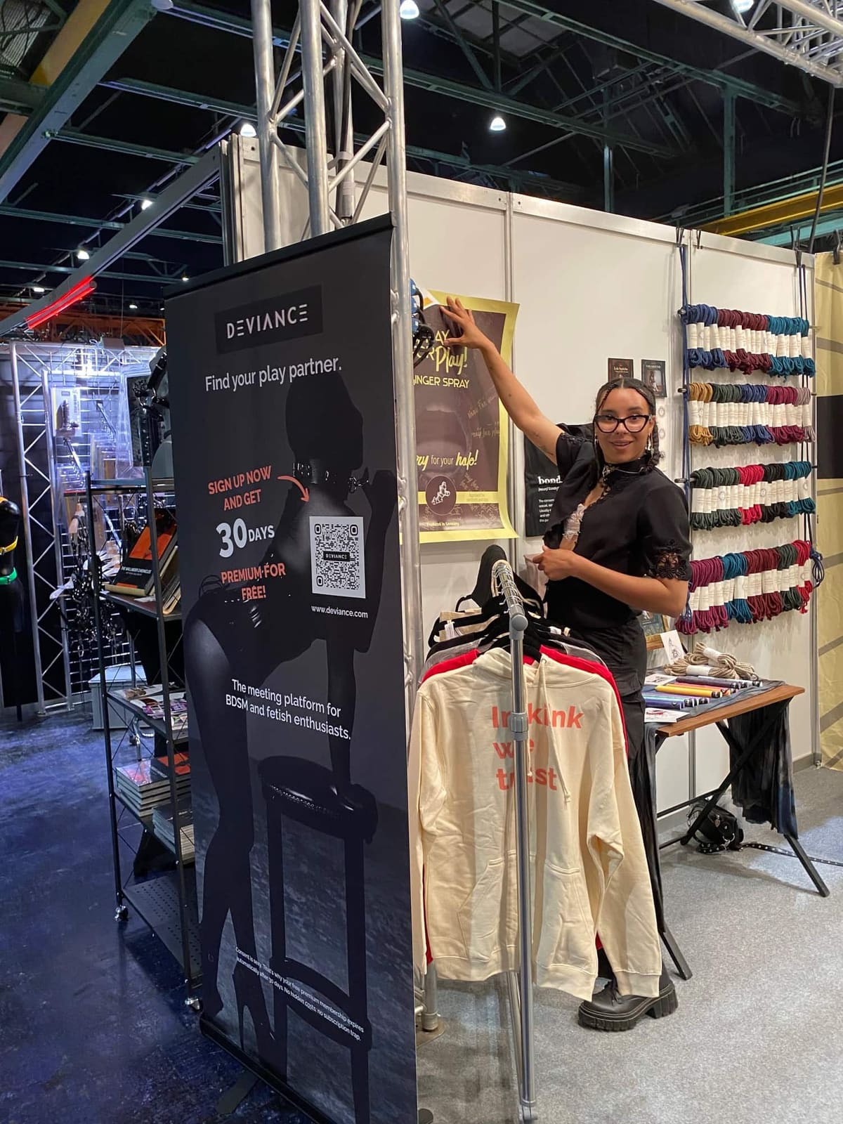 Persona en el estand de Deviance en BoundCon junto a un rollup y un perchero con sudaderas «In kink we trust», y una pared de cuerdas de bondage de colores a la derecha