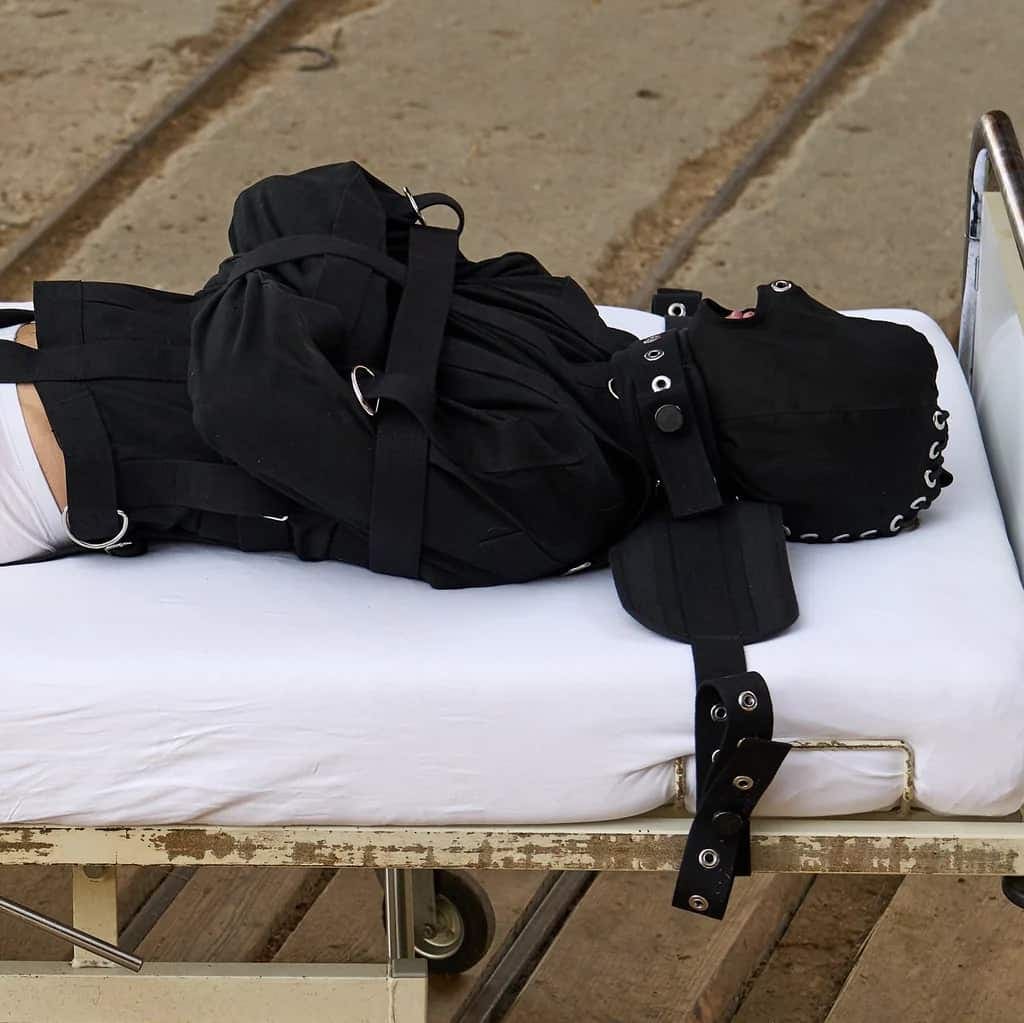 Persona con camisa de fuerza negra y máscara en la cabeza tumbada sobre una vieja camilla de hospital