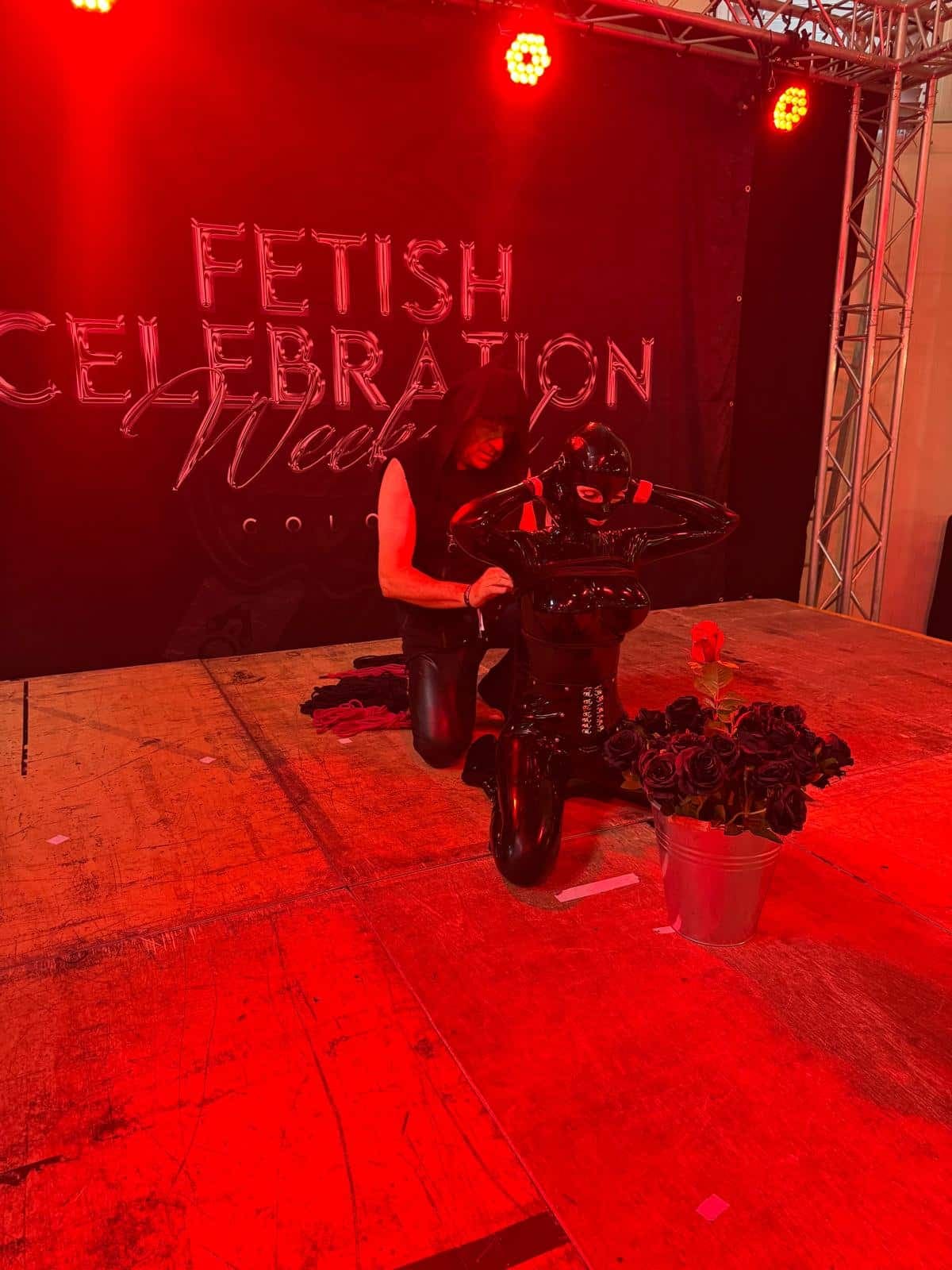 Escenario iluminado en rojo del Fetish Celebration Weekend: una persona arrodillada junto a otra con un traje completo de látex negro, y un cubo de rosas oscuras al lado