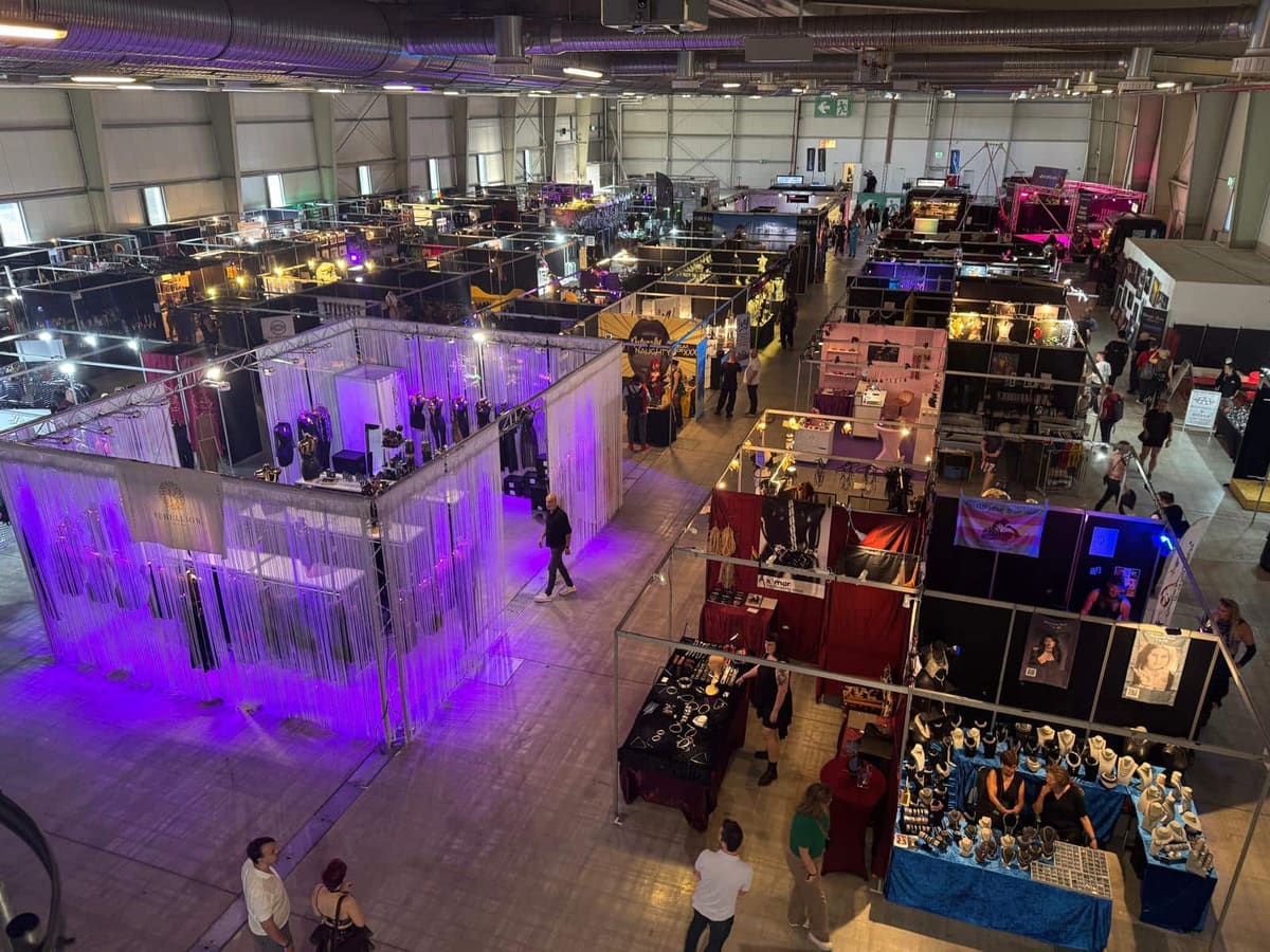 Vista desde arriba del pabellón de una feria fetichista con hileras de estands, cabinas iluminadas en violeta y visitantes en los pasillos