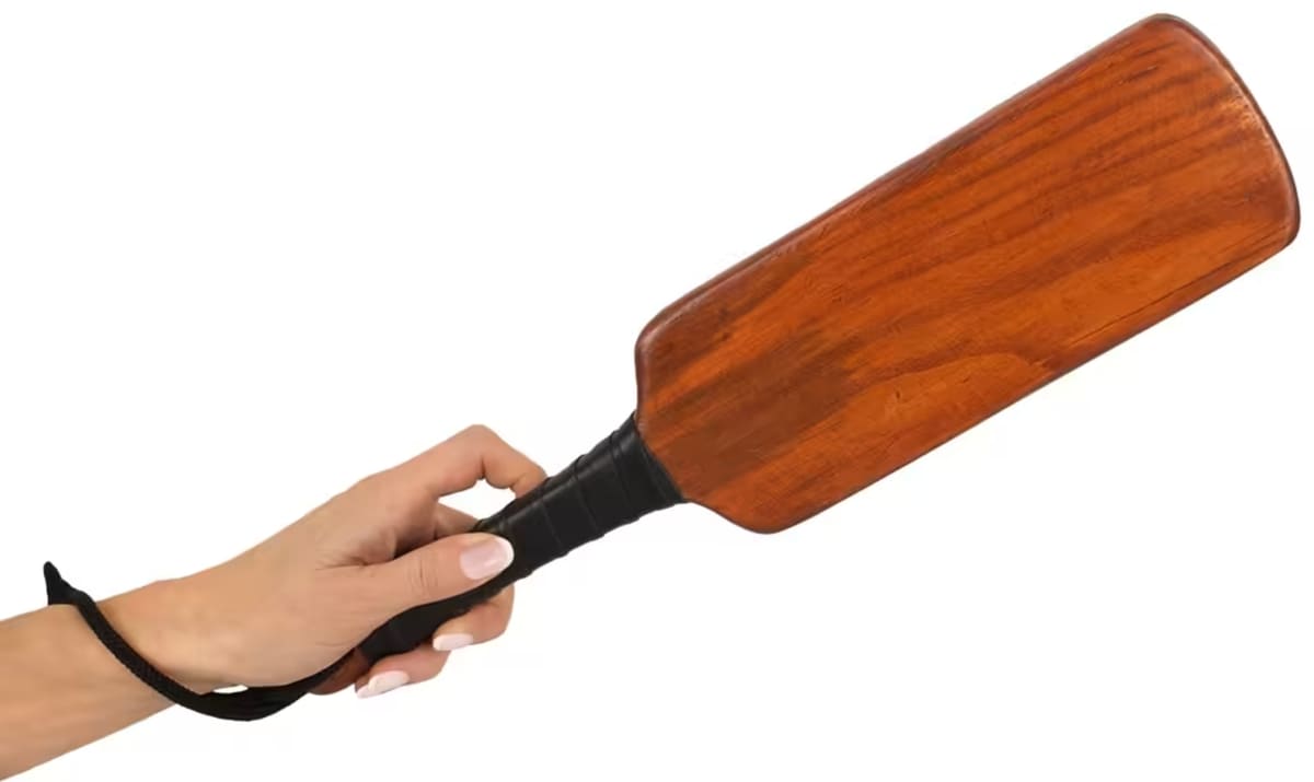 Una mano sostiene una gran pala de madera con mango envuelto en negro y lazo de muñeca sobre fondo blanco