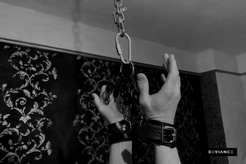 Fotografía en blanco y negro de dos manos con esposas de cuero sujetas con un mosquetón a una cadena del techo, con papel pintado barroco al fondo