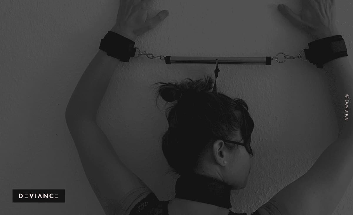 Fotografía en blanco y negro desde atrás: las muñecas de una persona separadas por muñequeras acolchadas en una barra sobre la cabeza