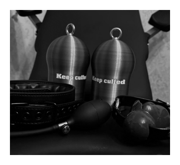 Fotografía en blanco y negro de un expositor con dos plugs metálicos grabados «Keep cuffed», un collar remachado y otros accesorios