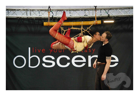 Suspensión de shibari en Obscene 2025 ante el cartel de la feria: una persona con traje rojo cuelga de una caña de bambú y besa al rigger