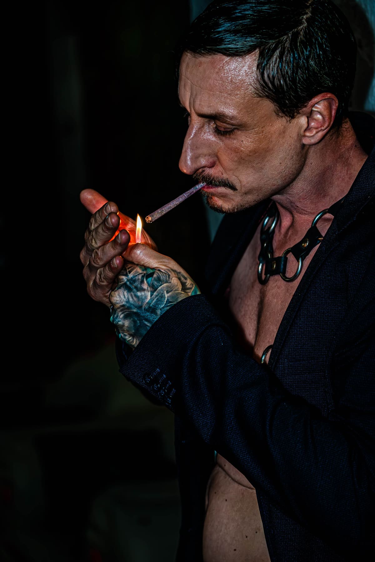 Fotografía nocturna en blanco y negro de una persona con americana abierta, collar de cuero y anillas en O encendiendo un cigarrillo