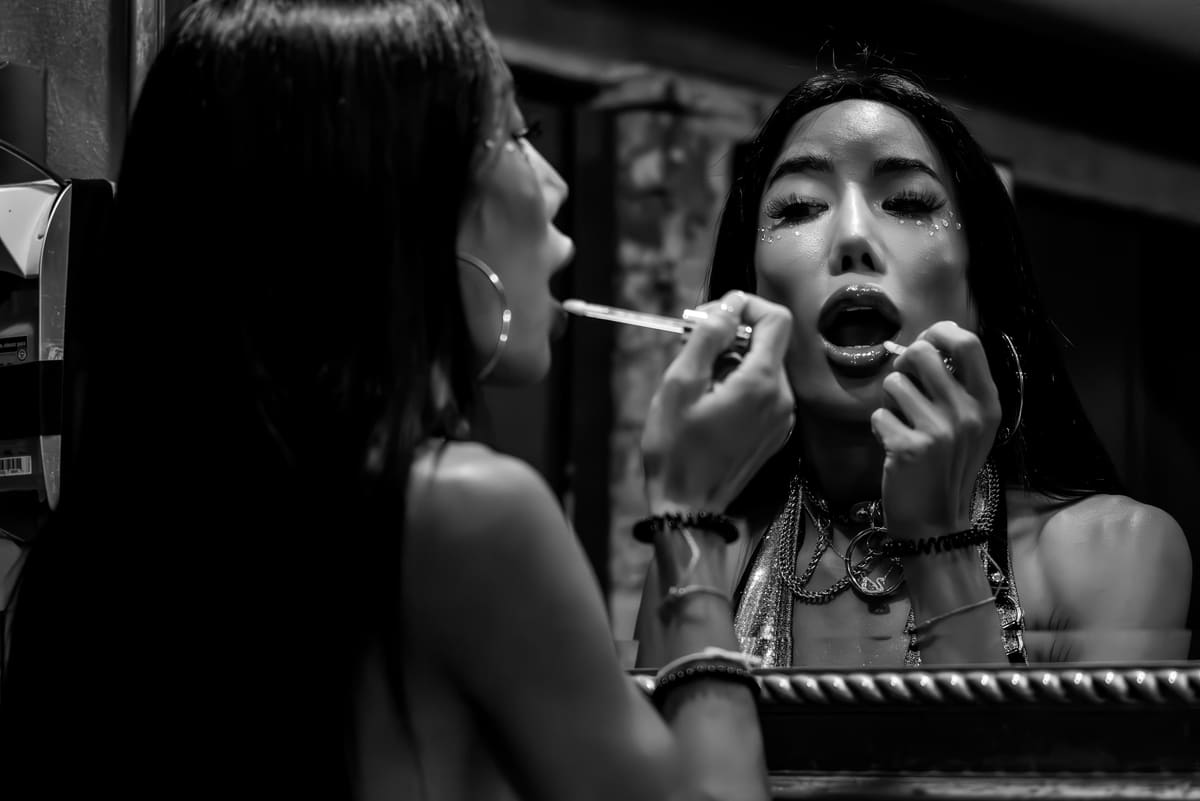 Fotografía en blanco y negro de una persona con purpurina en el rostro, cadenas y collar aplicándose brillo de labios ante un espejo