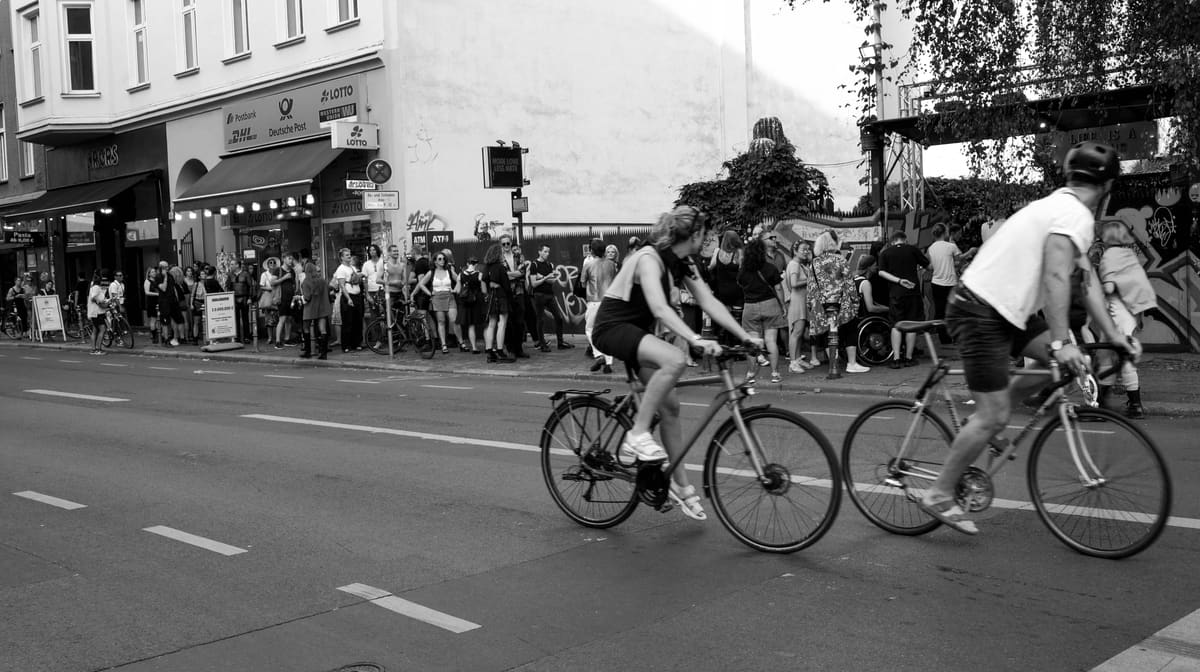 Escena callejera en blanco y negro: invitados esperan ante la entrada de la inauguración mientras pasan dos ciclistas