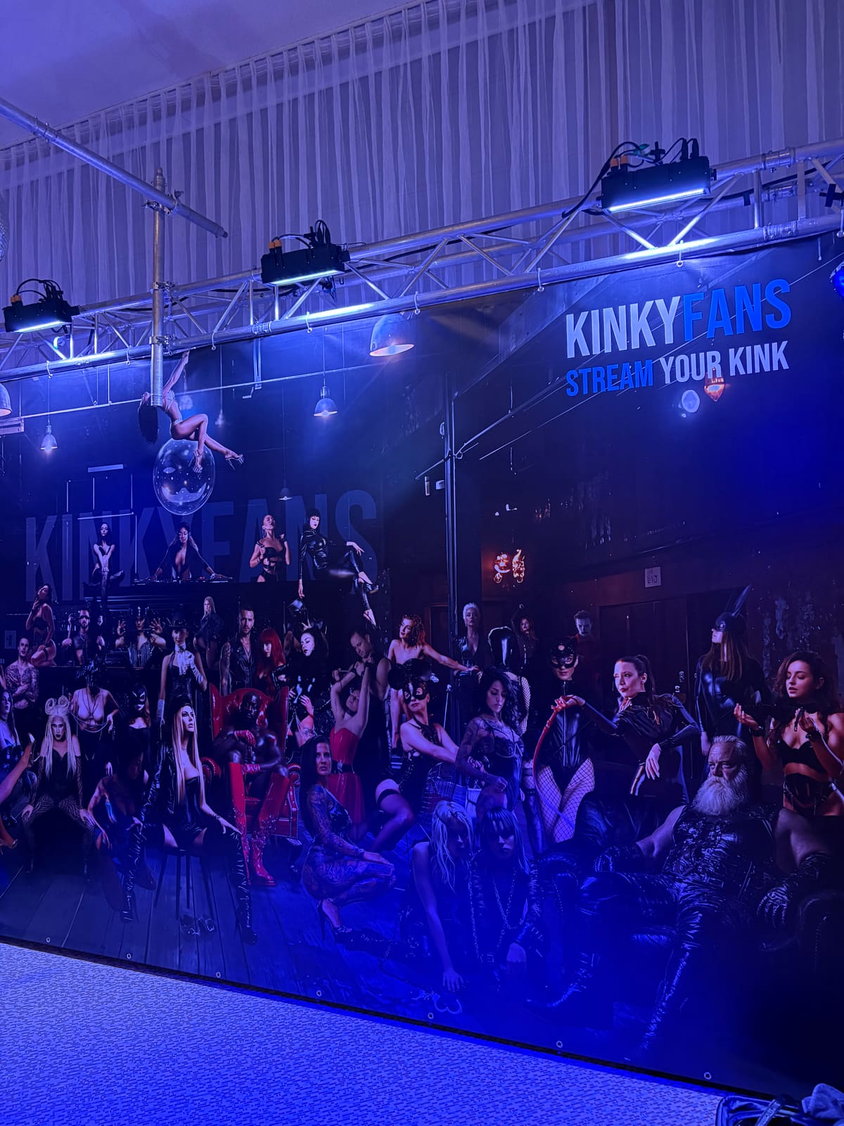 Escenario de KinkyFans iluminado en azul en Venus 2025 con el lema «Stream your kink» y un gran collage de artistas