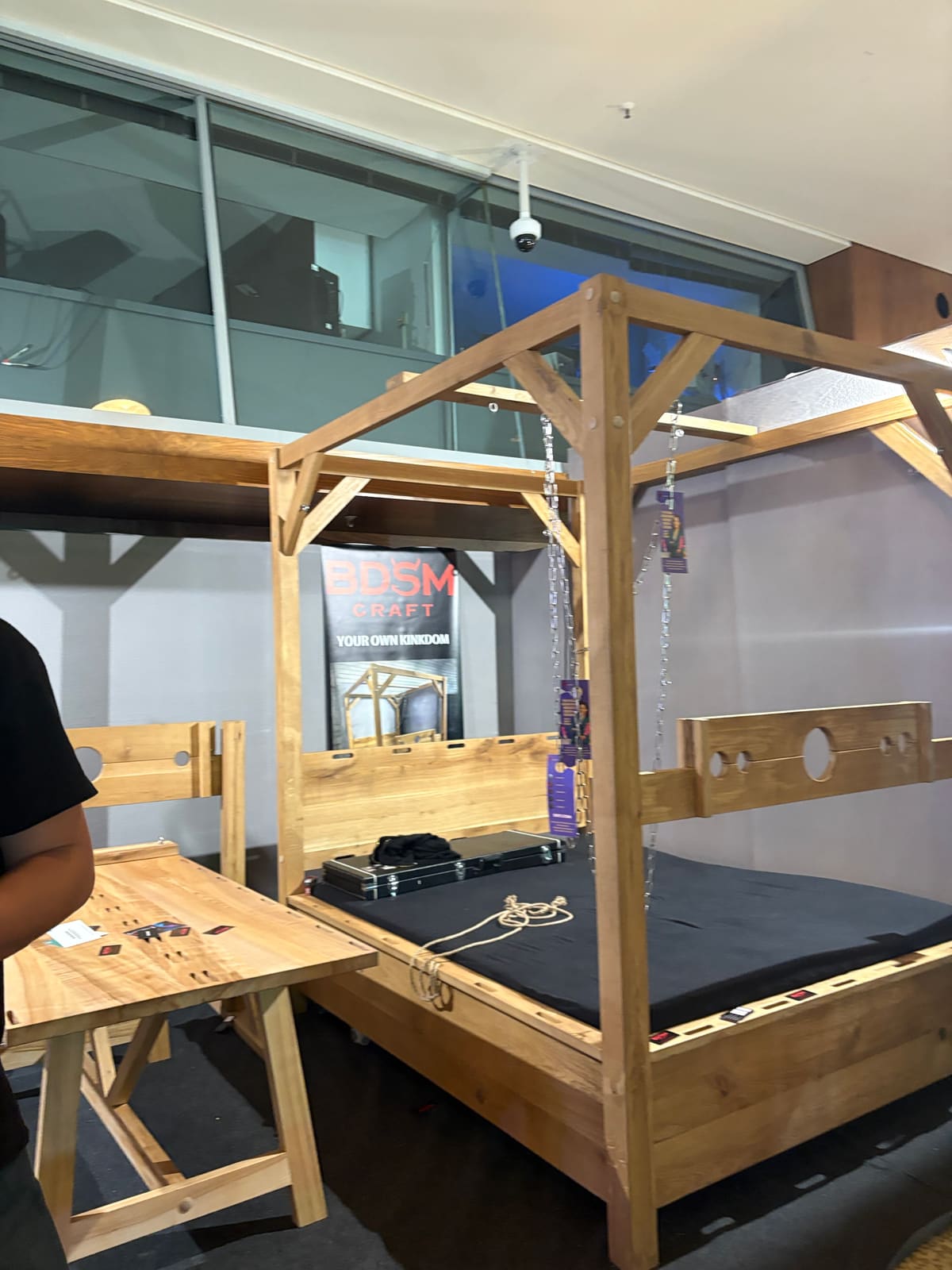 Estand de BDSM Craft en Venus 2025 con una cruz de San Andrés, un cepo y otros muebles de juego en madera clara