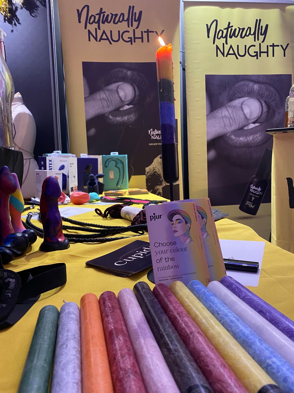 Estand de Naturally Naughty en Venus 2025: una vela multicolor encendida, filas de velas, consoladores y muestras de lubricante pjur sobre una mesa amarilla