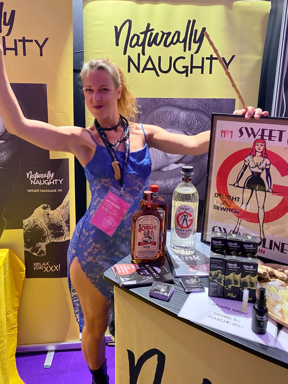 Persona con vestido azul de encaje y los brazos extendidos sosteniendo una vara en el estand de Naturally Naughty, con spray de jengibre, ginebra y folletos de Deviance en el mostrador