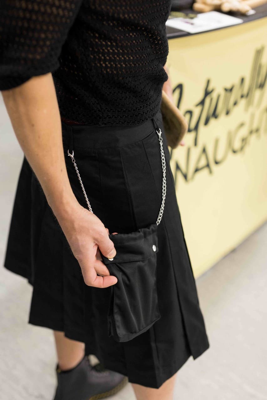 Detalle de una falda masculina negra con pliegues, bolsillo de parche y cadena plateada, con un cartel de Naturally Naughty detrás