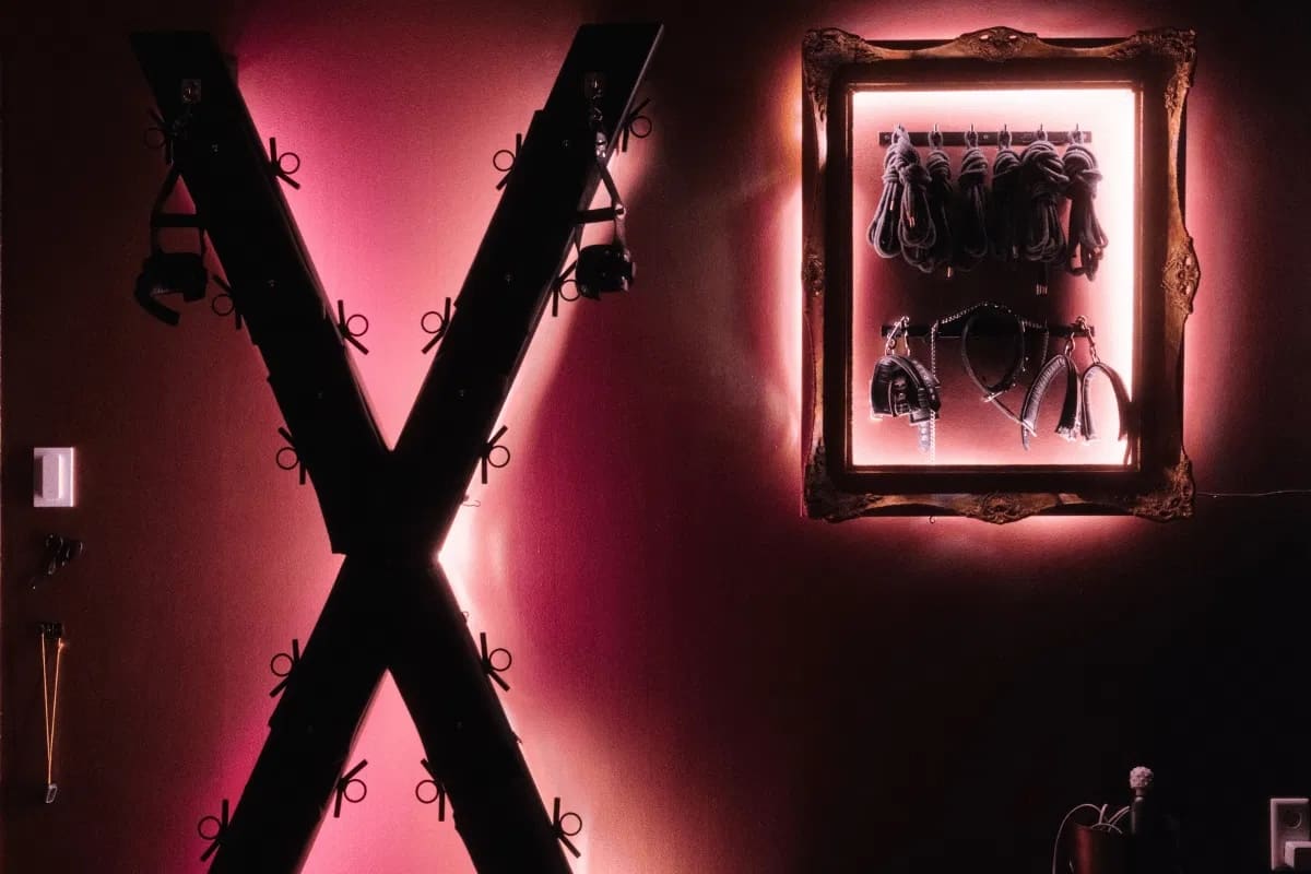 Sala de juego iluminada en rosa con una cruz de San Andrés en la pared y un marco con cuerdas, esposas y látigos