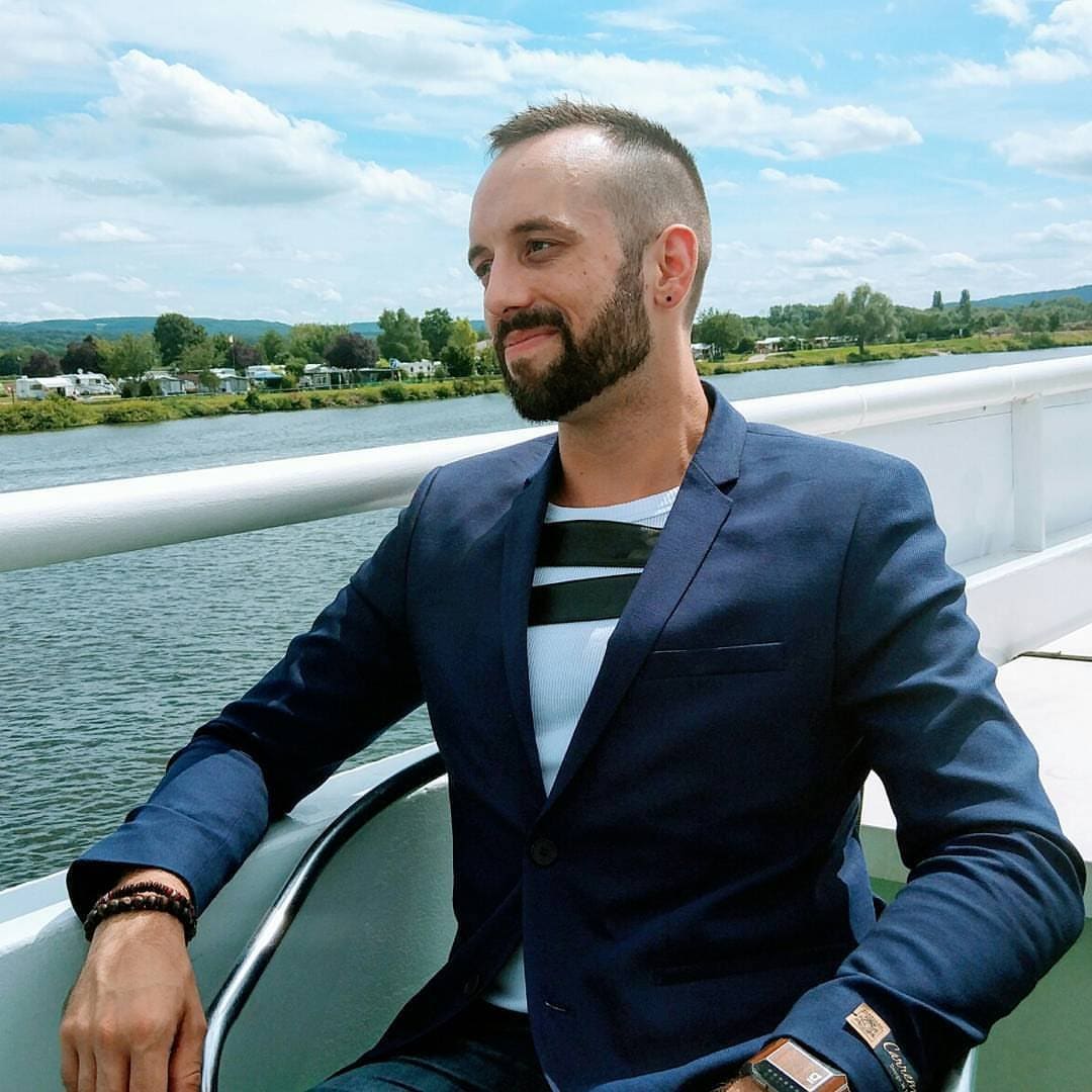 Jeff Mannes con americana azul sentado en la cubierta de un barco, con un río y una orilla verde al fondo
