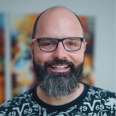 Retrato de un hombre sonriente con gafas, barba poblada y camiseta estampada ante un cuadro de colores